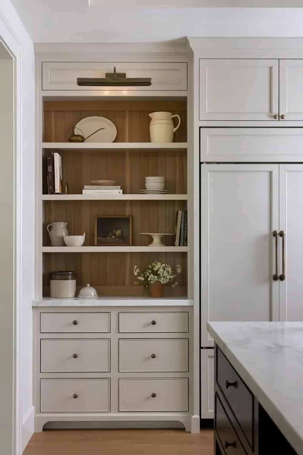 Kitchen open shelving niche with wood bead board back, styled ceramics, and brass picture light beside paneled refrigerator