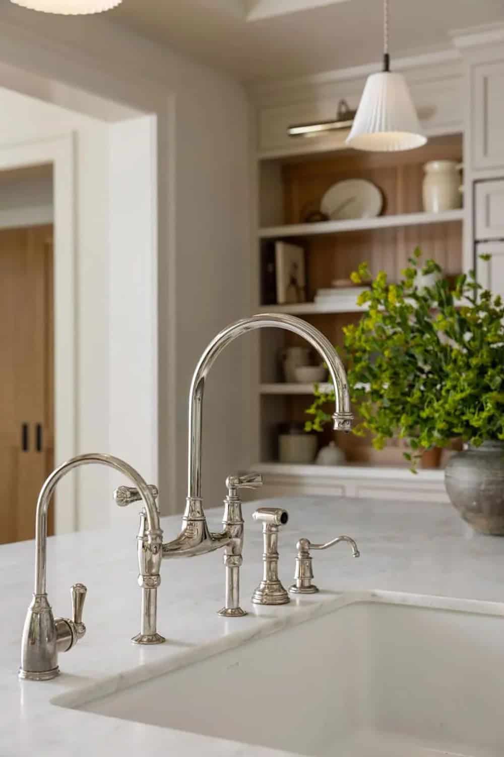 Close-up of polished nickel bridge faucet on white marble island with open kitchen shelving in background
