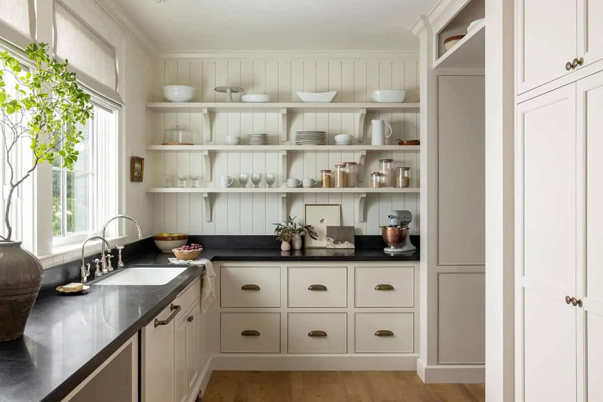 Butler's pantry with cream cabinets, black soapstone countertops, open white shelving, and farmhouse sink