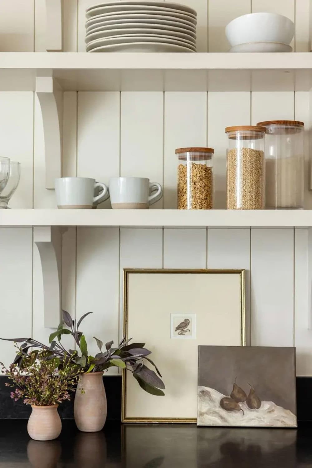 Close-up of butler's pantry open shelving with stacked plates, mugs, glass jars, and framed art on counter