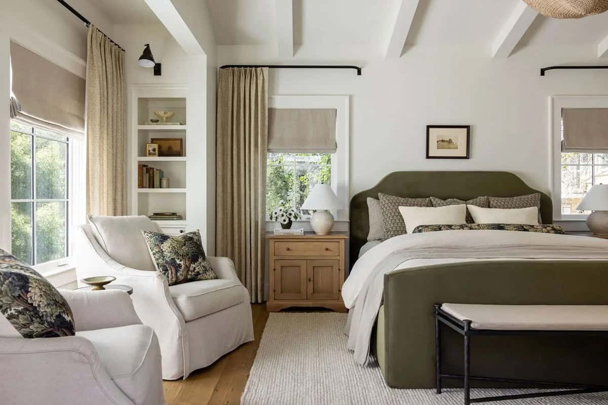 Primary bedroom with olive green upholstered bed, white armchairs, built-in shelves, and linen drapes