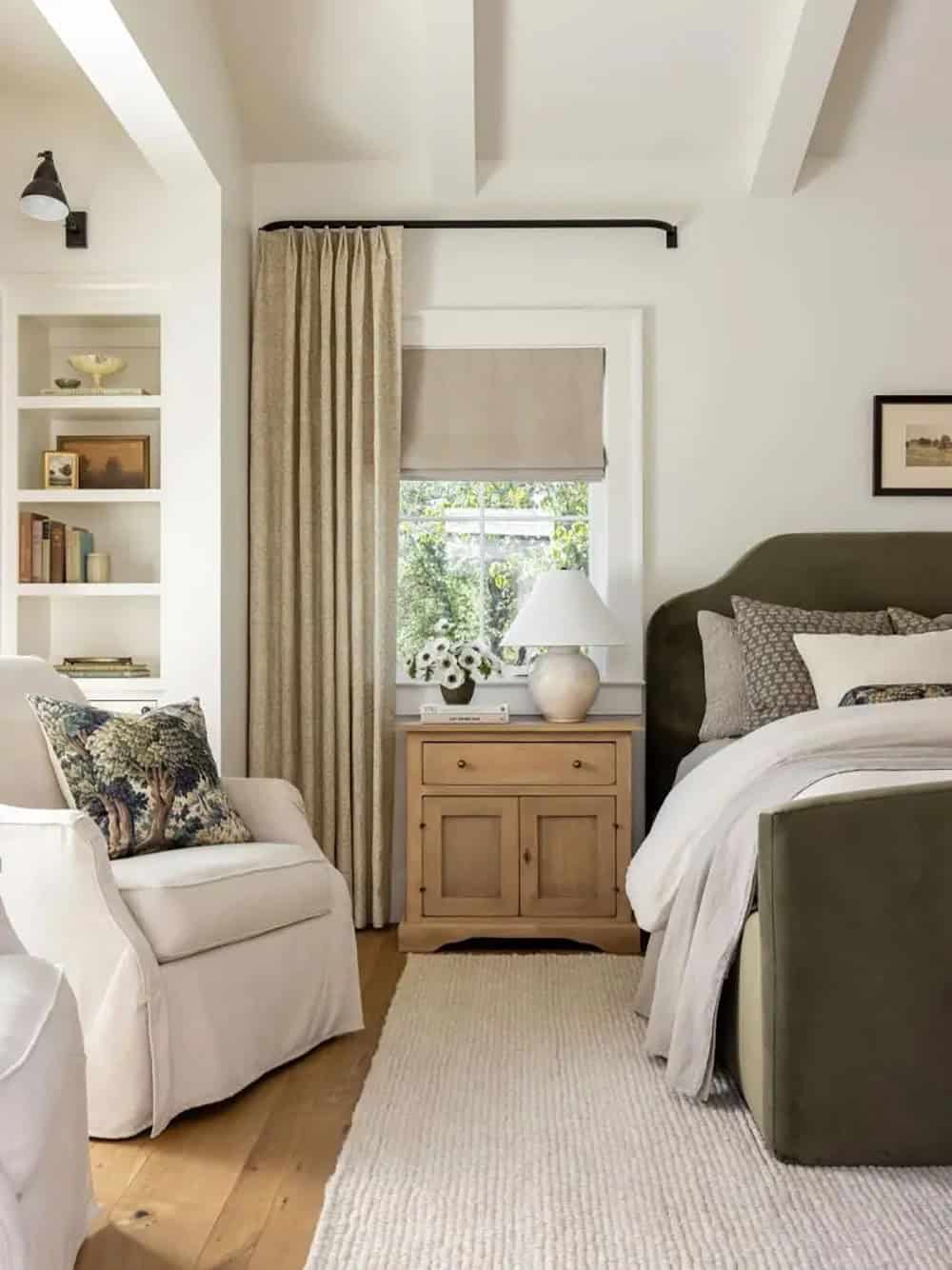 Primary bedroom with olive green upholstered bed, white armchair, antique nightstand, and linen curtains