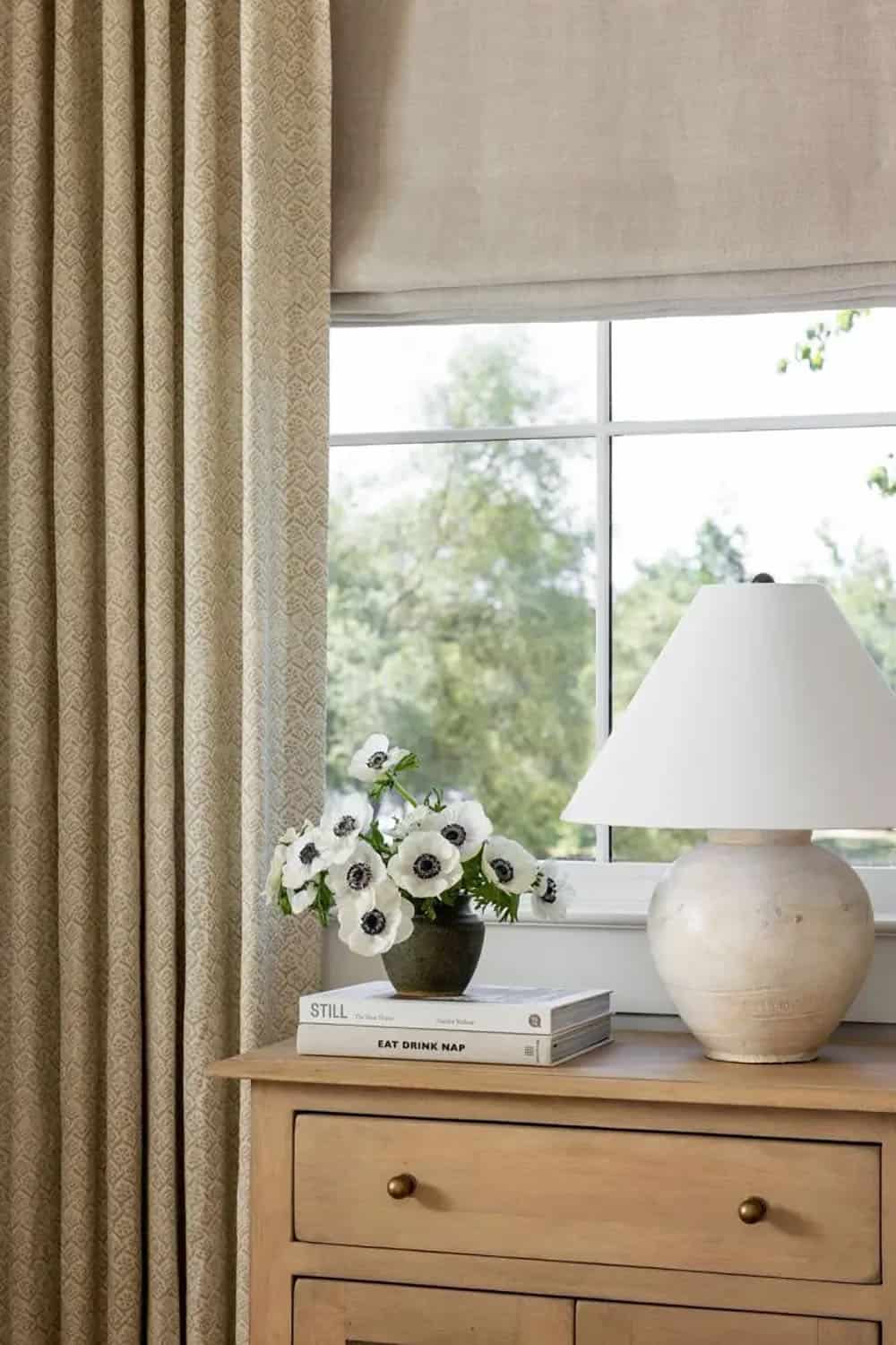 Close-up of bedroom nightstand with white anemone flowers, ceramic lamp, and patterned linen drapes
