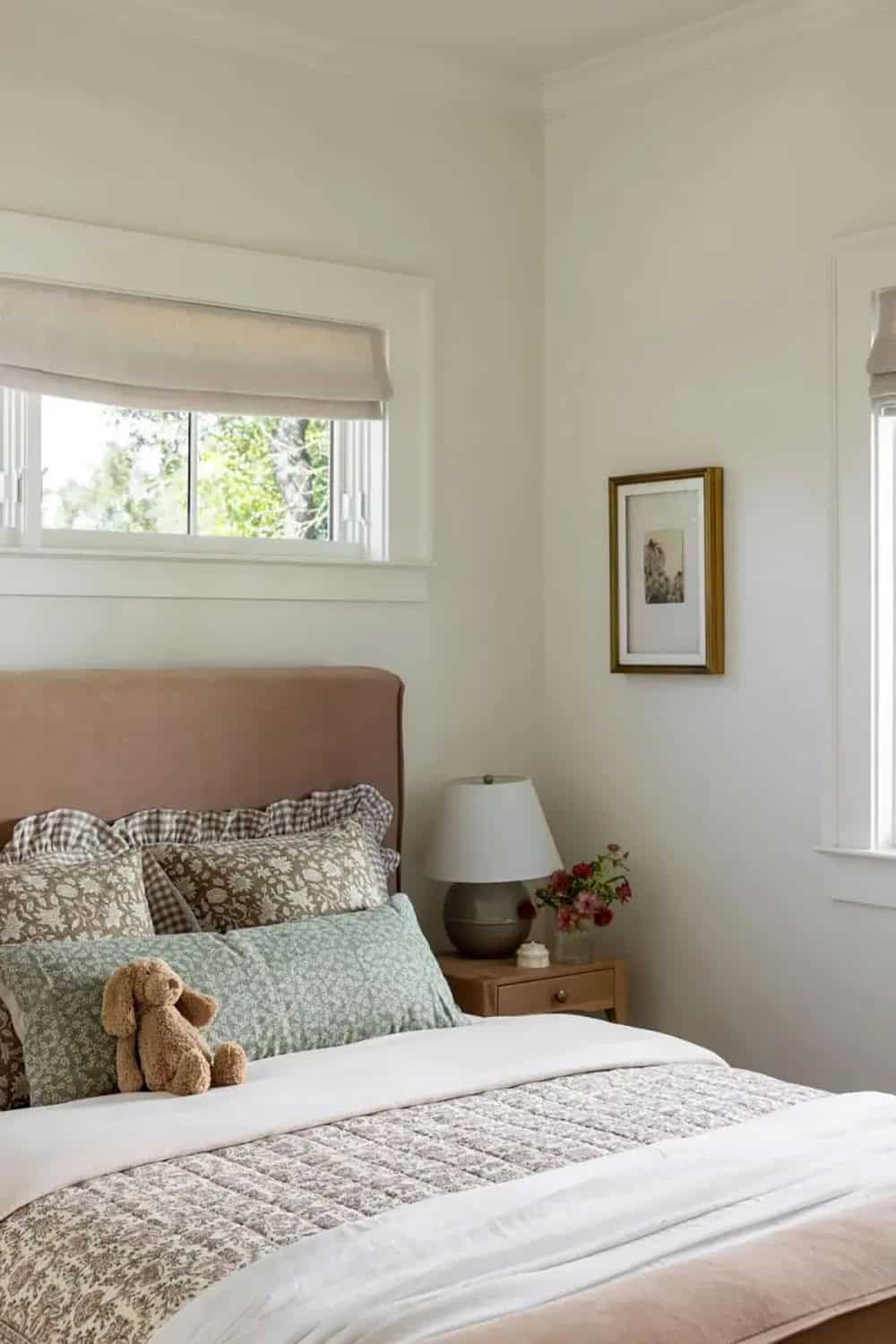 Kid's bedroom with blush upholstered headboard, floral and gingham bedding, teddy bear, and ceramic lamp