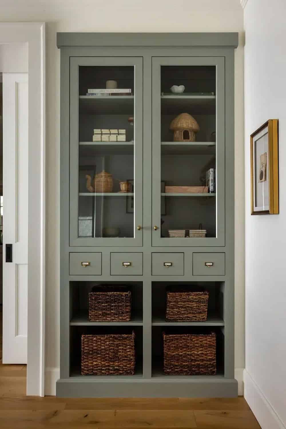 Sage green built-in cabinet with glass doors, brass hardware, and wicker storage baskets