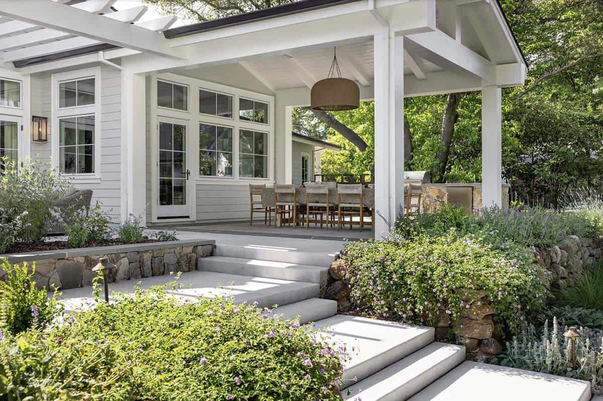 Covered outdoor dining porch with stone steps, wicker pendant, and blooming garden beds