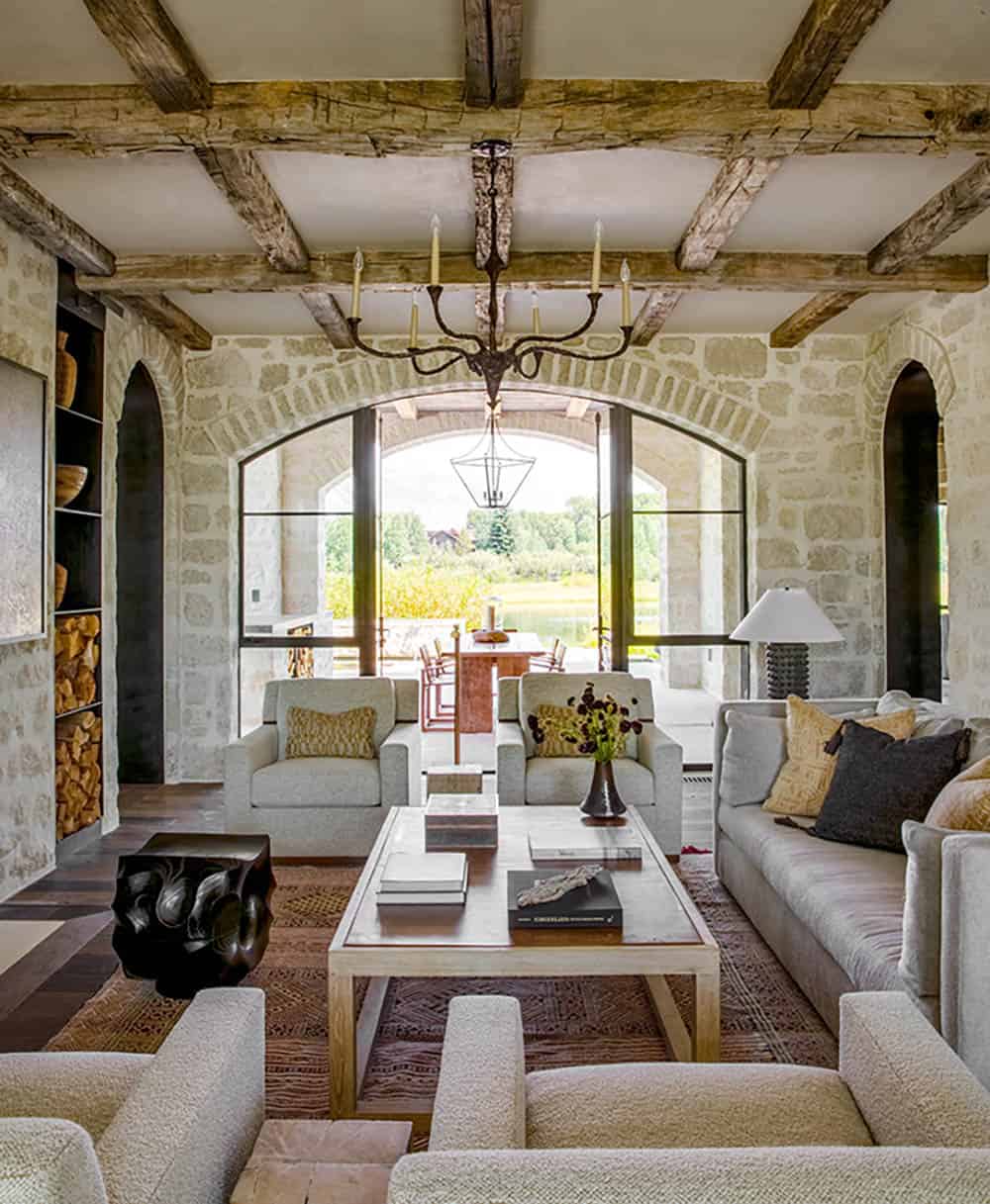 Stone living room with reclaimed beam ceiling, iron chandelier, and arched window to outdoor dining
