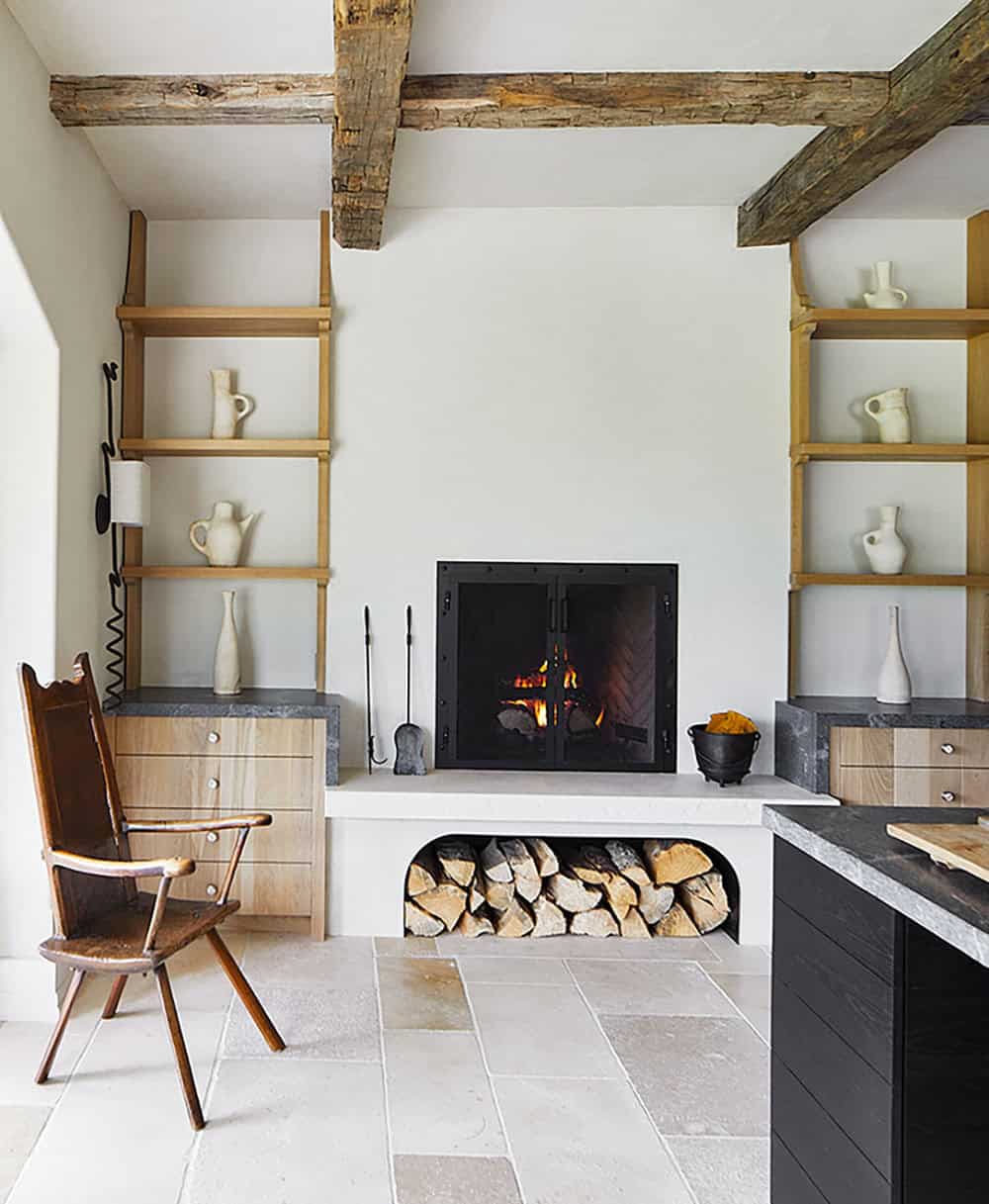 Kitchen fireplace with open wood storage, oak ladder shelving, white ceramic vessels, and antique chair