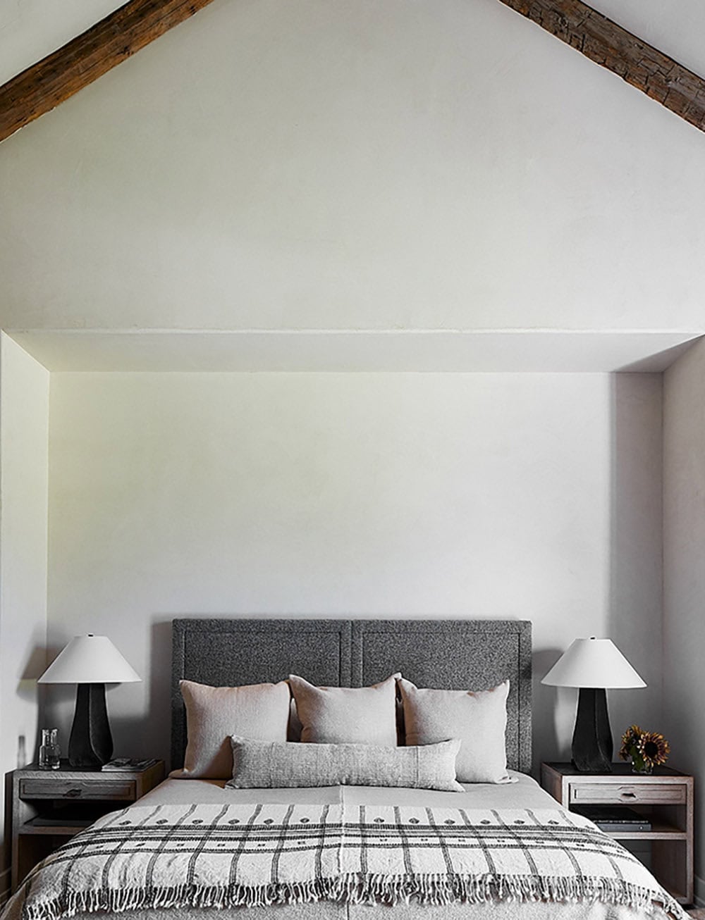 Minimalist bedroom with vaulted ceiling, gray upholstered headboard, plaid coverlet, and matching table lamps