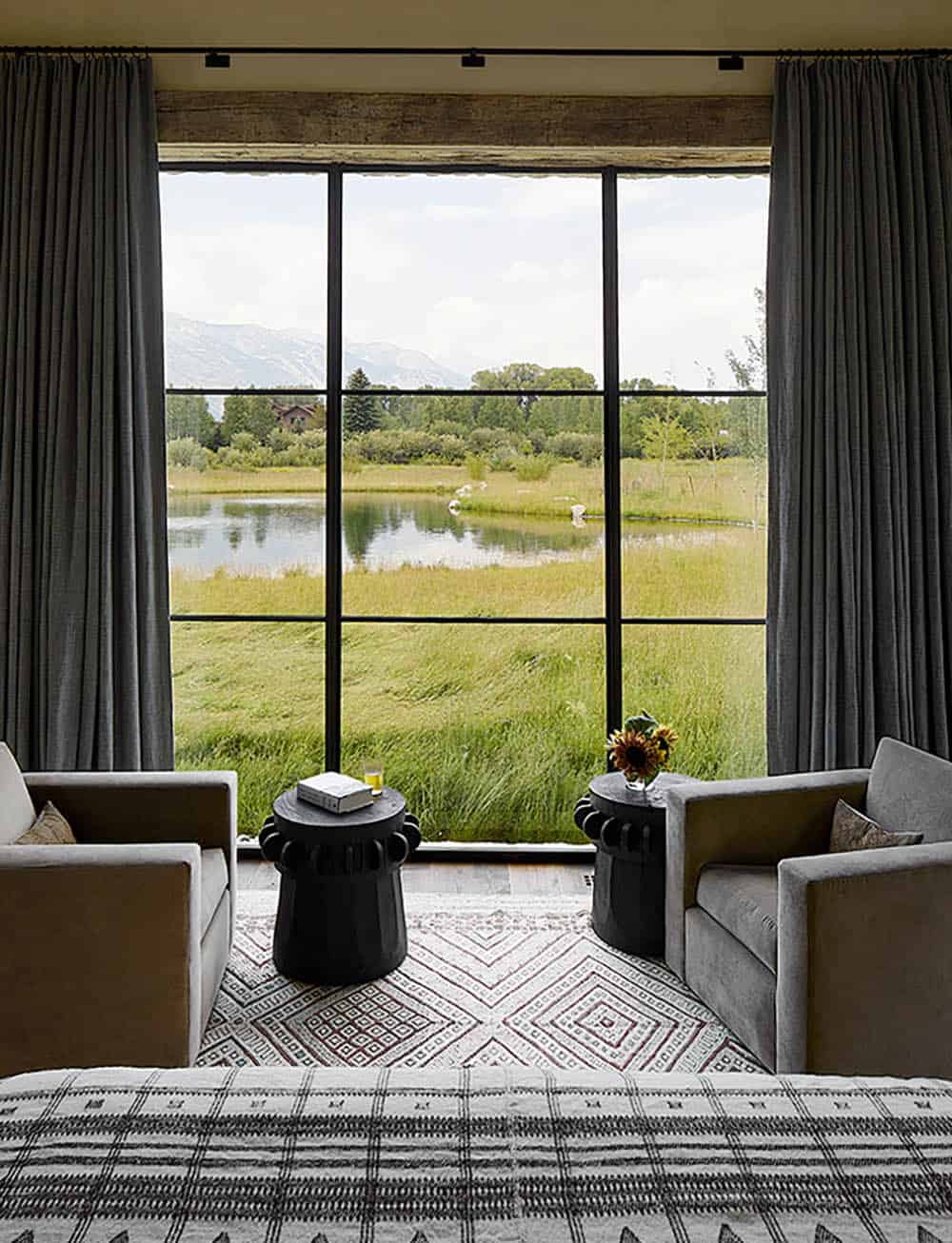 Bedroom sitting area with gray armchairs, black pedestal side tables, and panoramic pond and mountain views