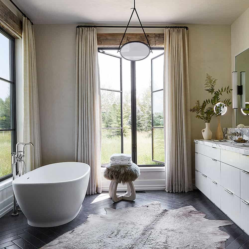 Primary bathroom with freestanding white soaking tub, herringbone black tile floor, cowhide rug, and garden views