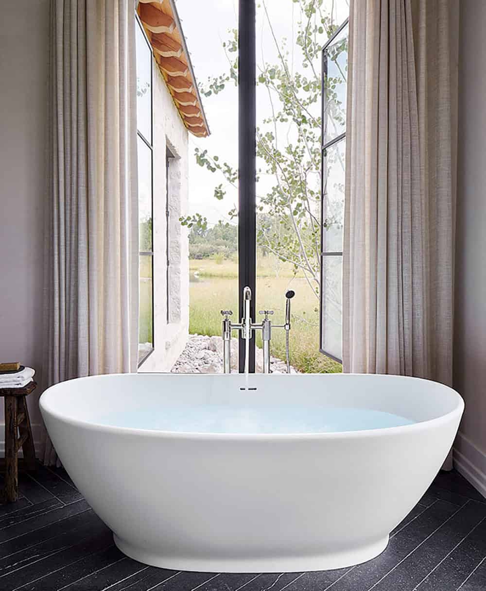 Freestanding white oval soaking tub beside floor-to-ceiling steel window overlooking wetland meadow