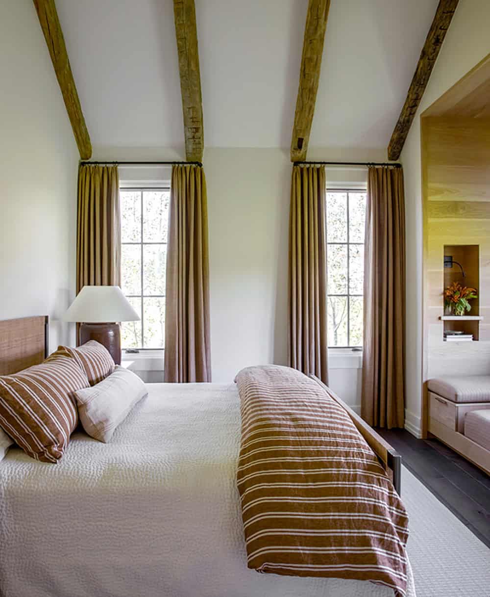 Guest bedroom with ochre striped linen coverlet, warm curtains, reclaimed beams, and wood-paneled reading nook