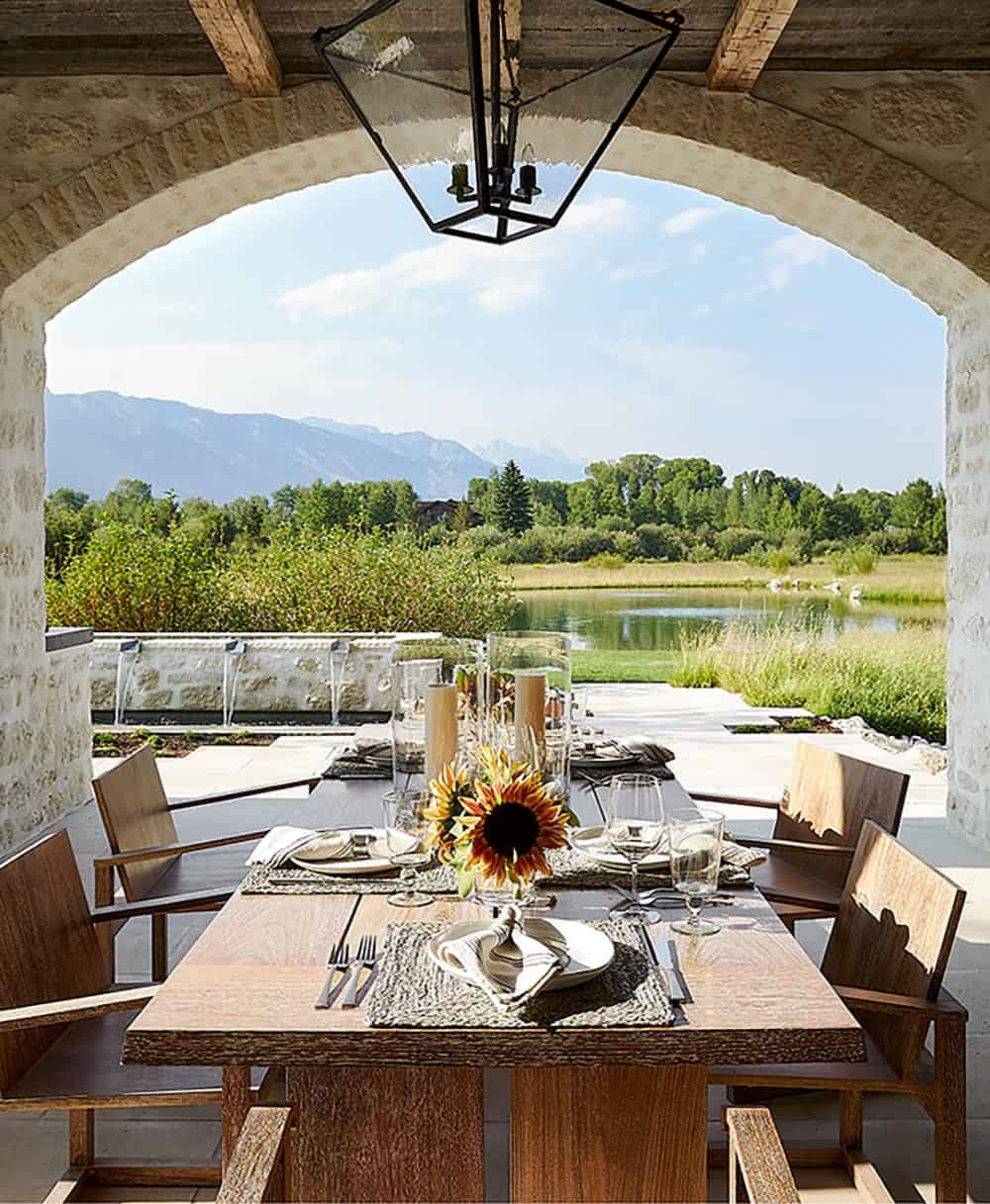 Covered outdoor dining under stone arch with set table, sunflowers, and Jackson Hole mountain panorama
