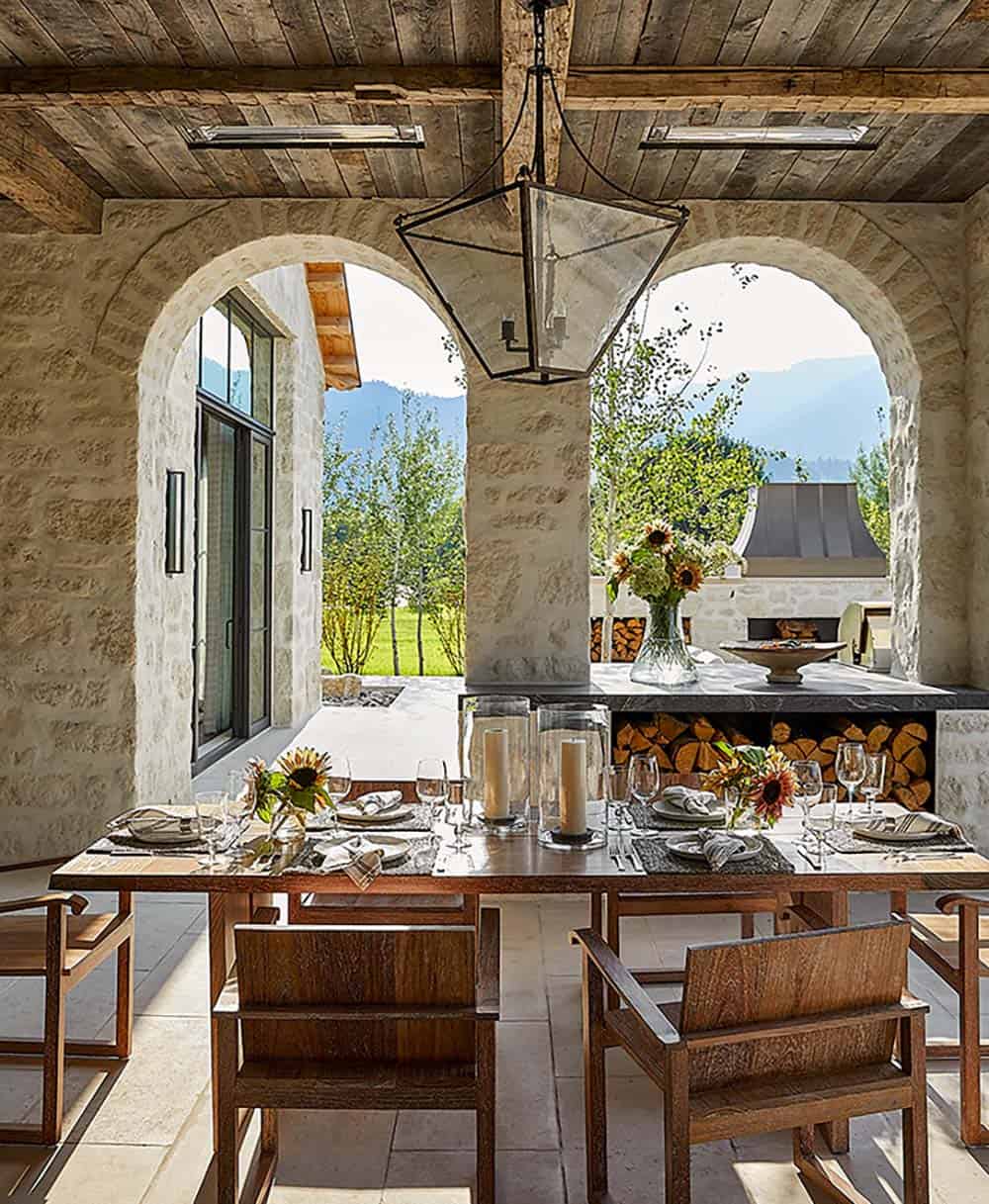 Stone-arched outdoor loggia with reclaimed wood ceiling, long dining table, and outdoor kitchen beyond