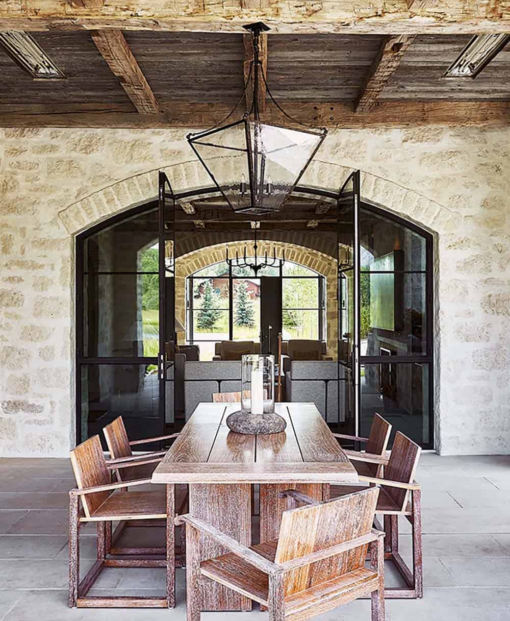 Covered outdoor dining area with weathered wood table, iron lantern pendant, and arched glass doors to interior