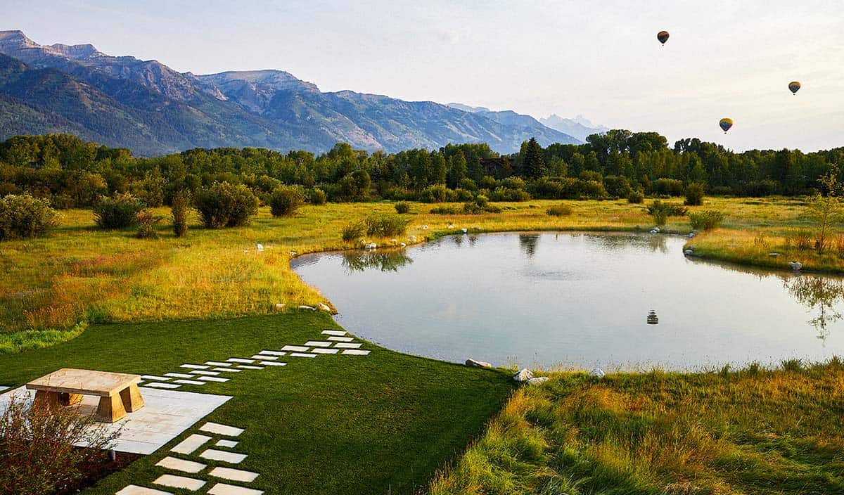 Aerial view of property pond with stepping stone path, wildflower meadow, and hot air balloons over Tetons