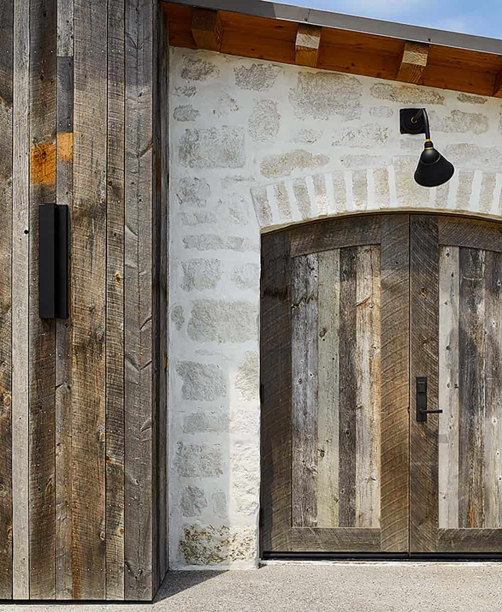 Arched stone entry with reclaimed wood double doors, black sconce, and vertical barn wood siding