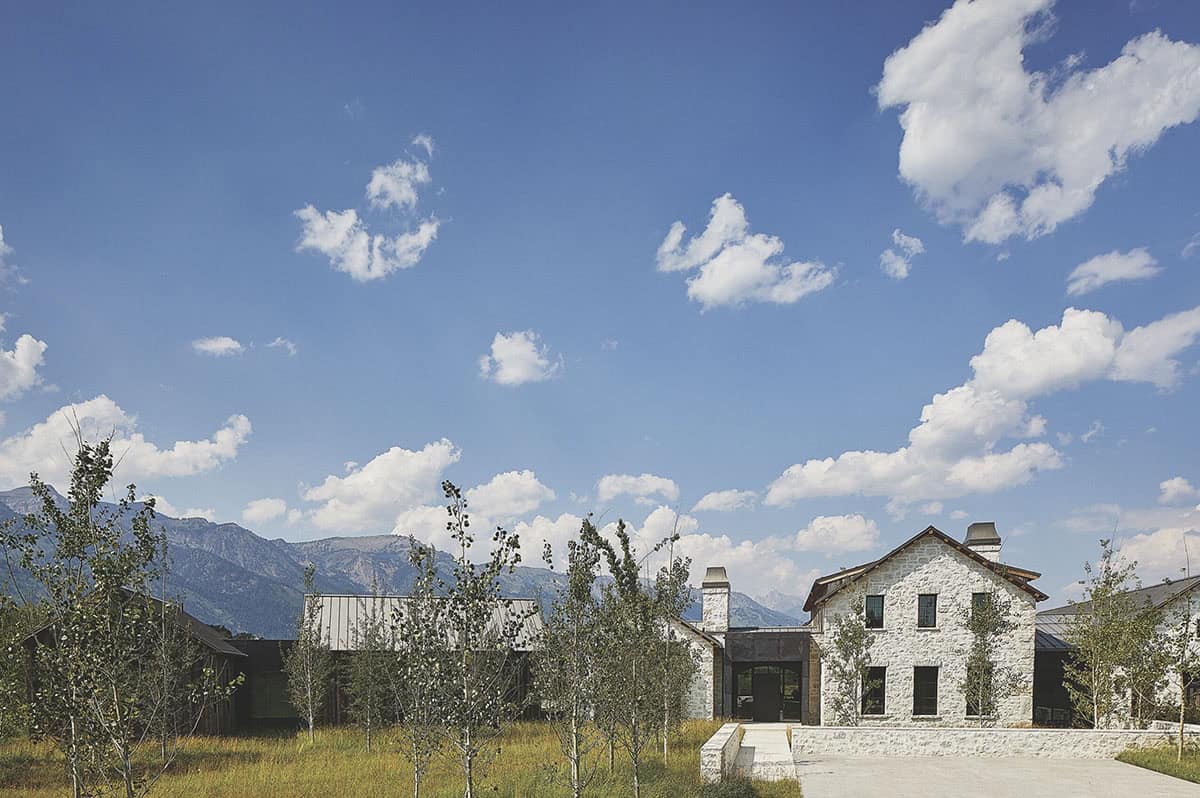 Front exterior of stone mountain home with metal roof, chimneys, young aspen grove, and Teton range backdrop