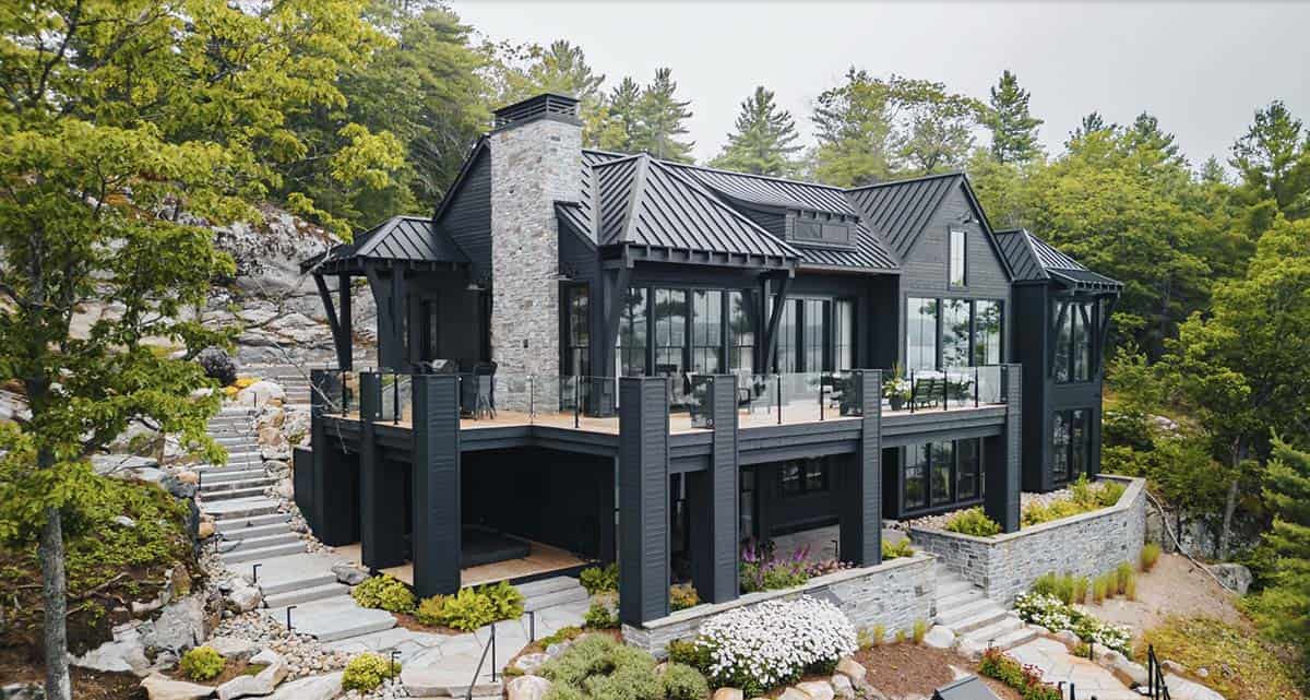 Aerial view of dark charcoal cottage with metal roof, stone chimney, and wrap-around deck nestled among trees