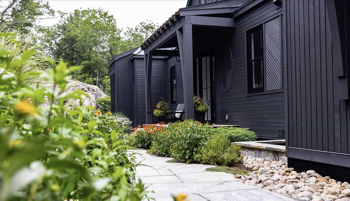 Dark-painted cottage entrance with stone pathway, colorful garden beds, and covered porch