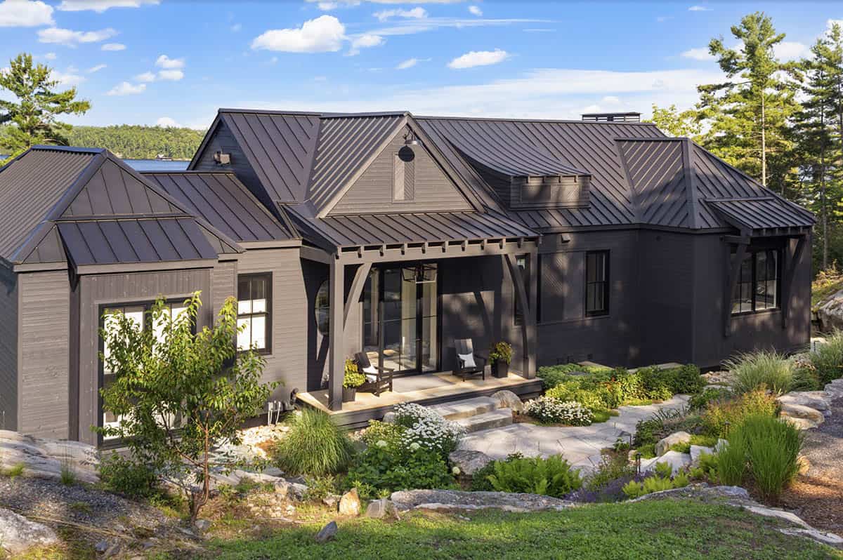 Dark grey lakeside cottage with complex metal roofline, covered porch, and naturalistic rock garden