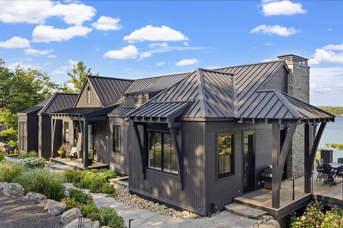 Dark cottage with dramatic multi-gabled metal roof, stone chimney, and lakeside deck