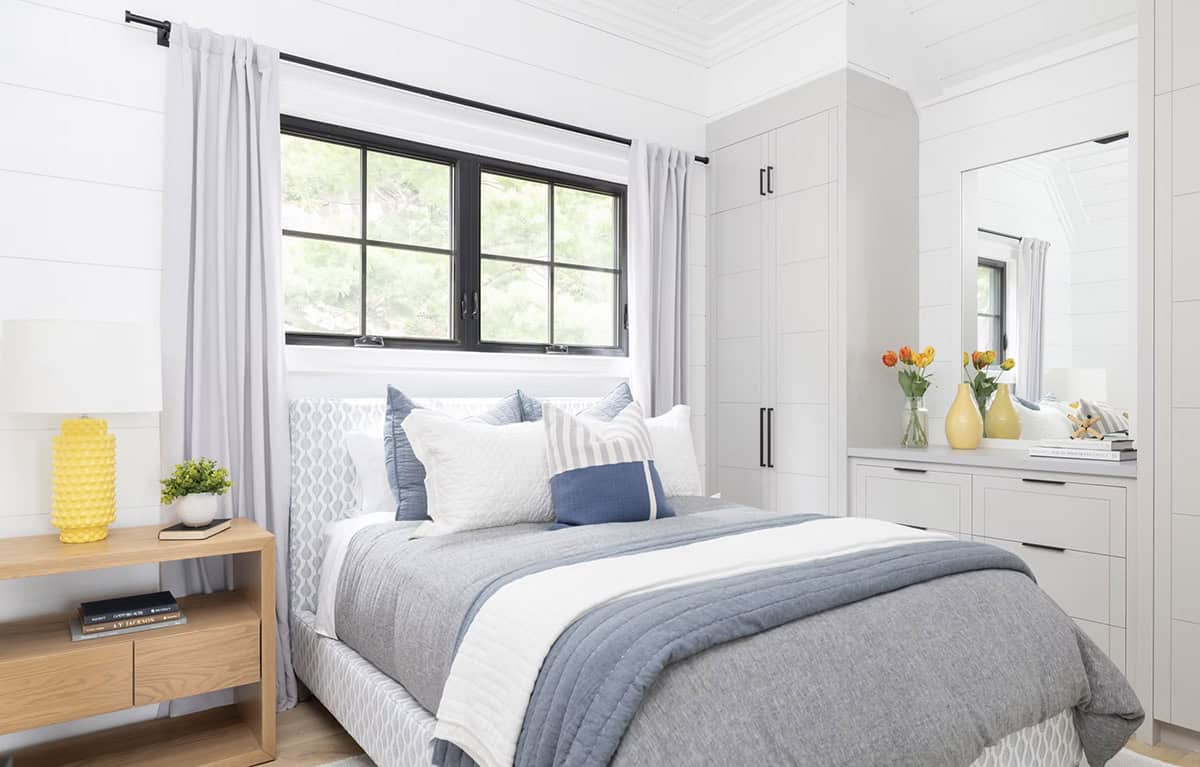Boathouse bedroom with blue and white bedding, yellow lamp, black-framed windows, and built-in gray wardrobes