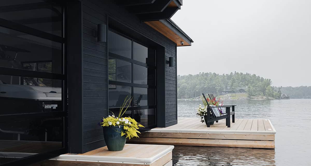Boathouse exterior with black board-and-batten siding, glass garage door, and lakeside Adirondack chairs