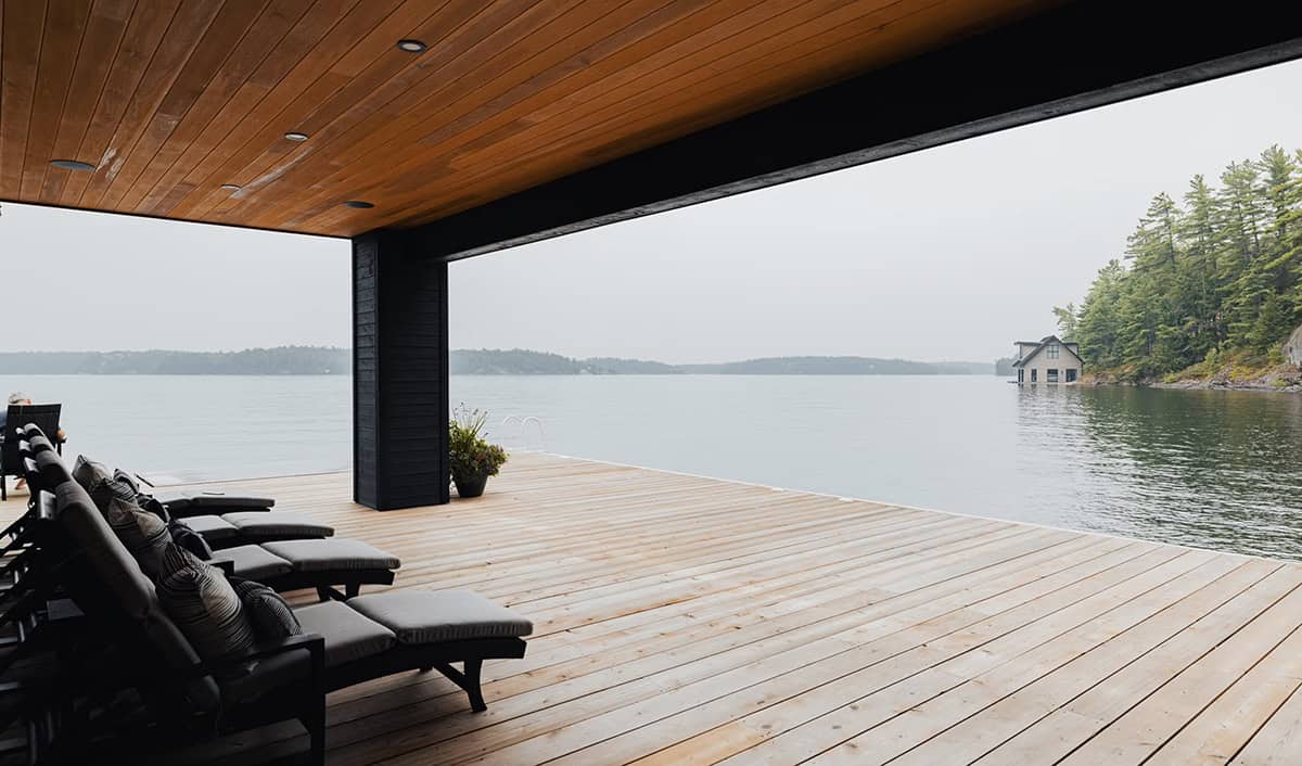 Covered boathouse dock with lounge chairs overlooking calm lake and distant boathouse