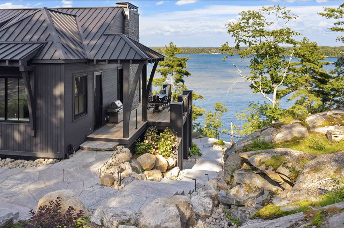 Side view of dark cottage with flagstone terracing, boulder landscaping, and blue lake vista