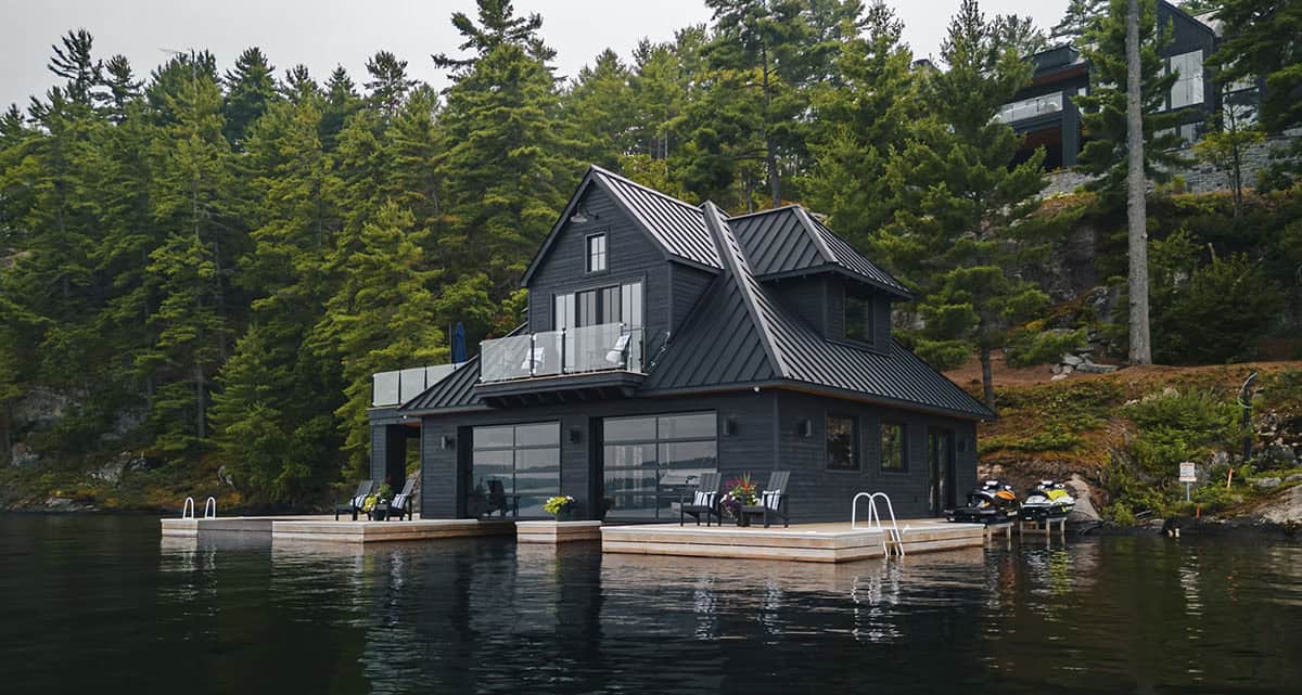 Black boathouse on Lake Joseph with standing-seam metal roof, glass garage doors, cedar docks, and Adirondack chairs