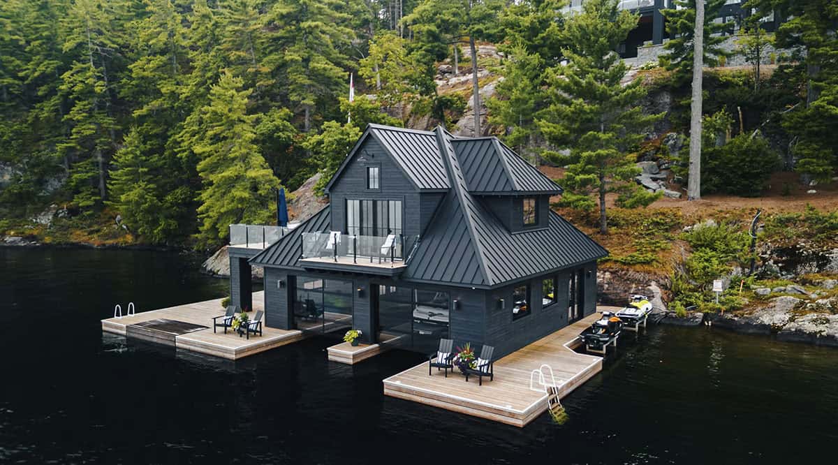 Aerial view of black boathouse with multi-gabled metal roof, cedar docks, Adirondack chairs, and jet skis