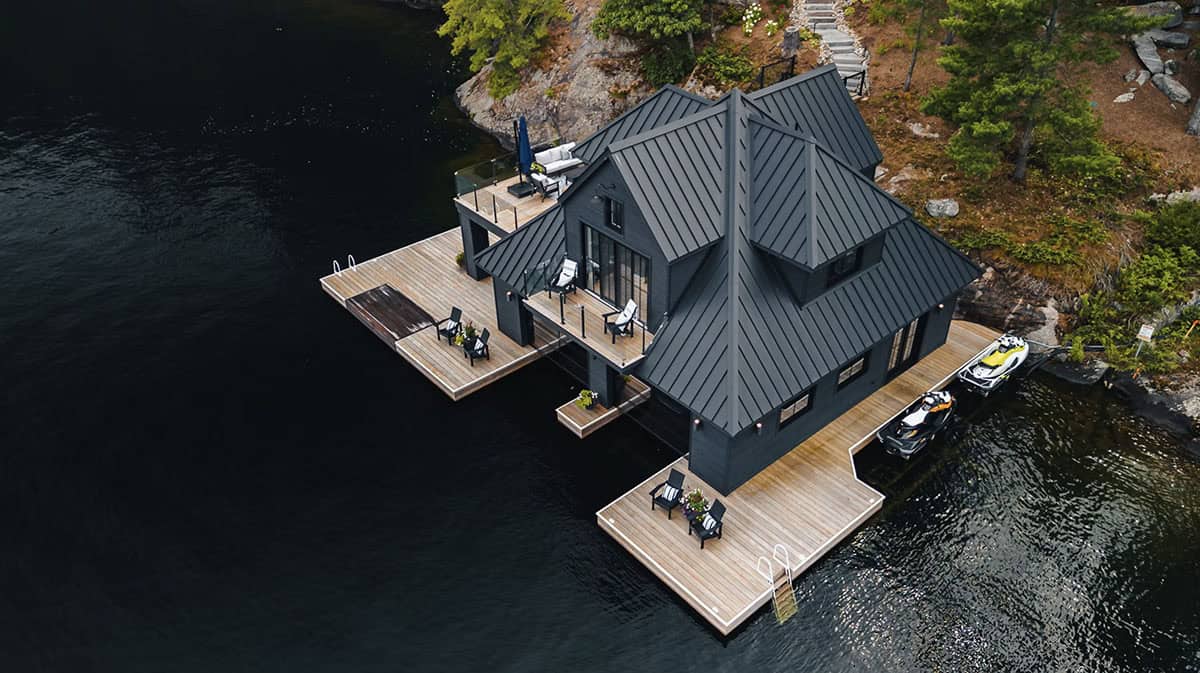 Elevated aerial view of black boathouse with cedar decks, rooftop patio, and jet skis on dark lake water