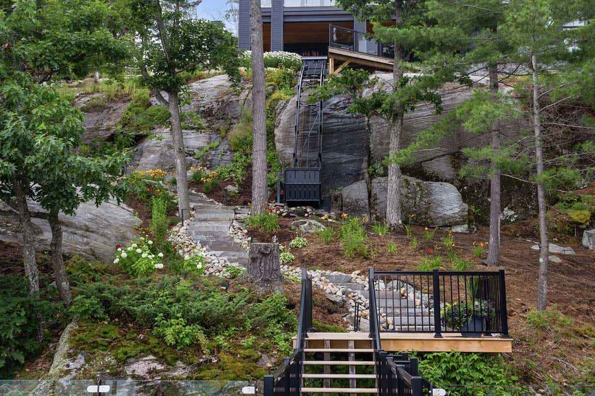 Steep Canadian Shield hillside with black steel funicular, stone steps, wildflower gardens, and pine trees connecting cottage to waterfront