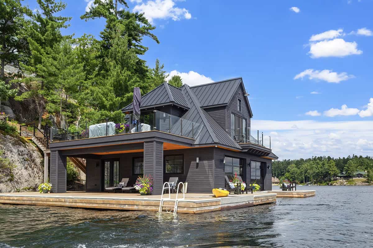 Black boathouse on Lake Joseph with multi-gabled metal roof, upper rooftop terrace, covered lower deck, and cedar docks on a sunny summer day
