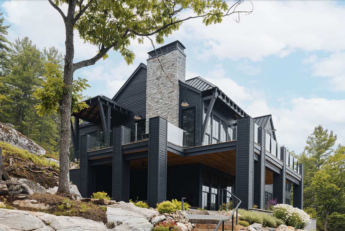 Dramatic view of dark cottage lake-facing facade with glass railings and exposed wood ceiling soffit