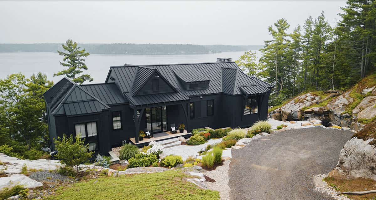 Aerial view of all-black cottage with complex roofline perched on rocky lakeside terrain
