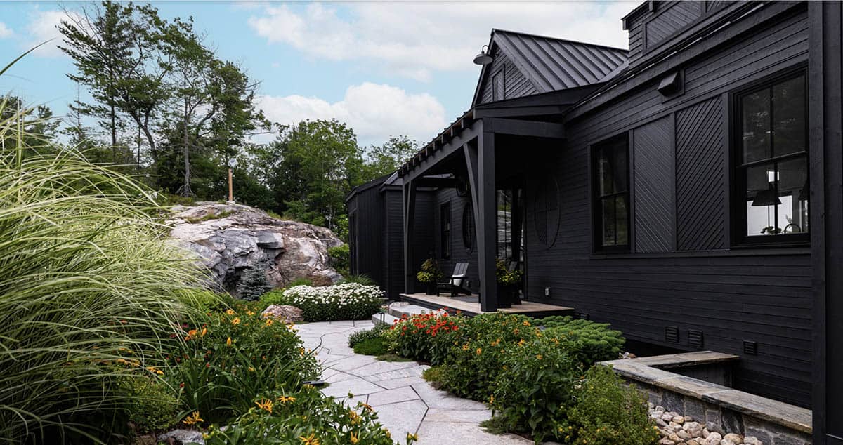 Black cottage side elevation with flagstone walkway, mixed perennial garden, and exposed granite outcrop