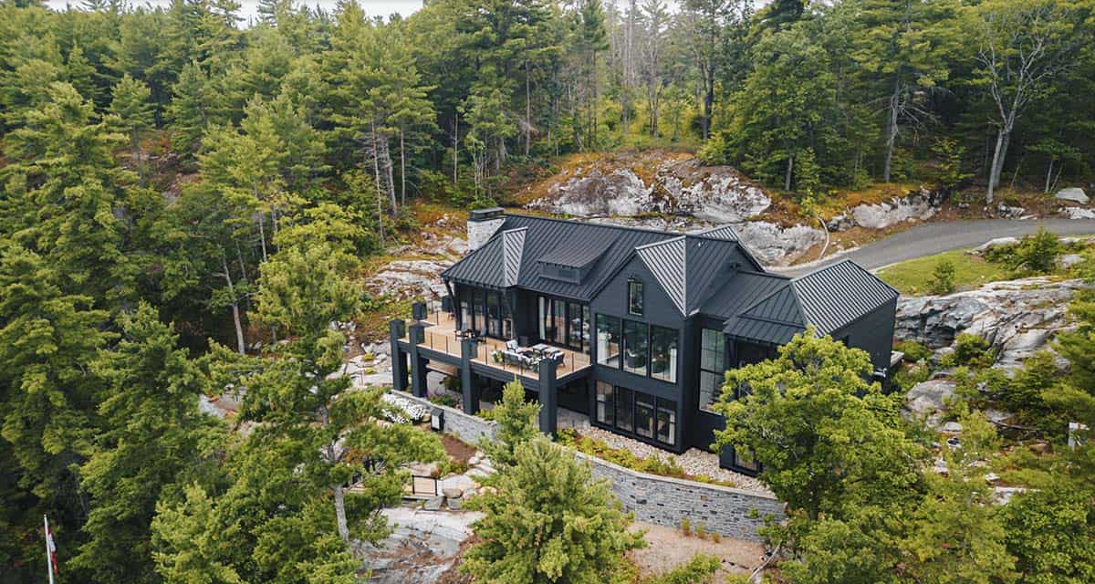 Aerial view of dark two-story cottage with large deck surrounded by autumn-tinted forest