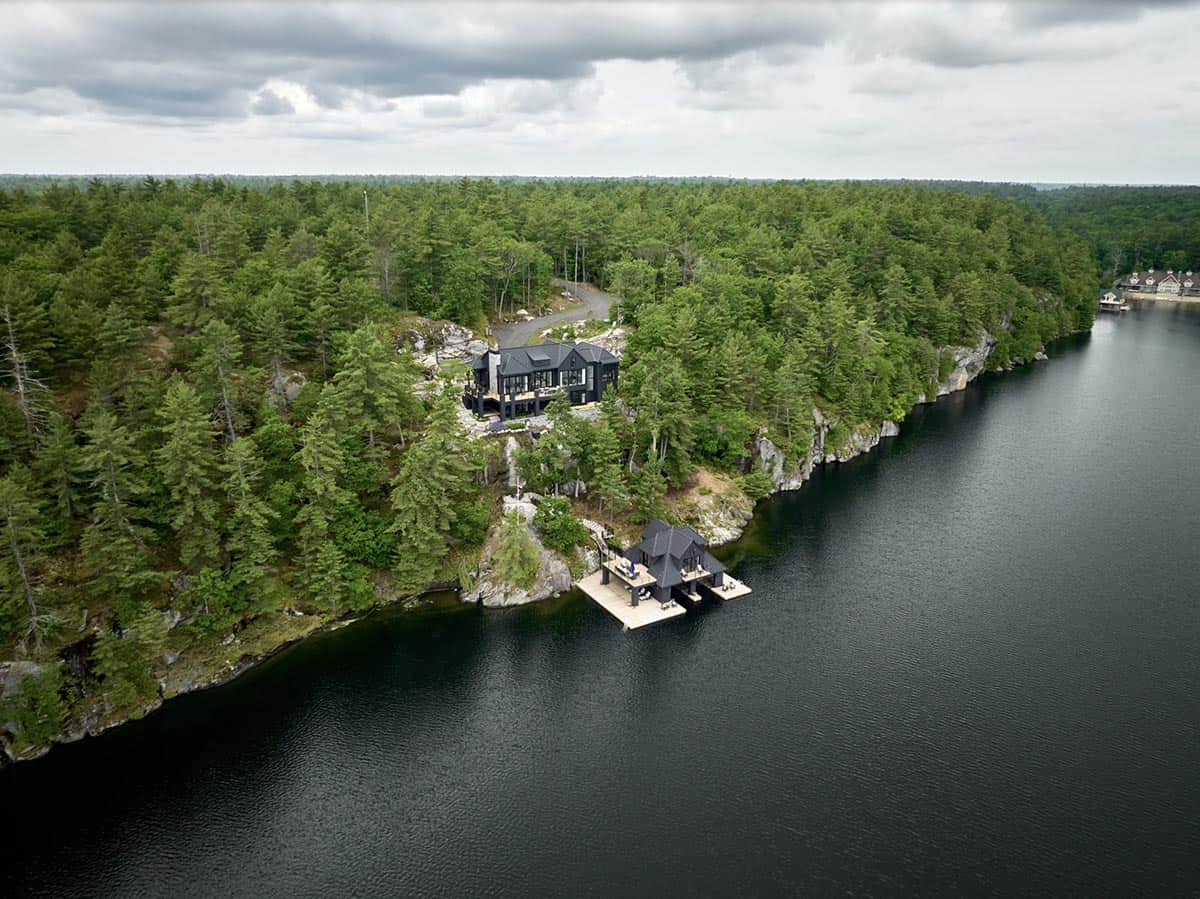 Wide aerial of black cottage and boathouse on forested rocky peninsula surrounded by dark lake water