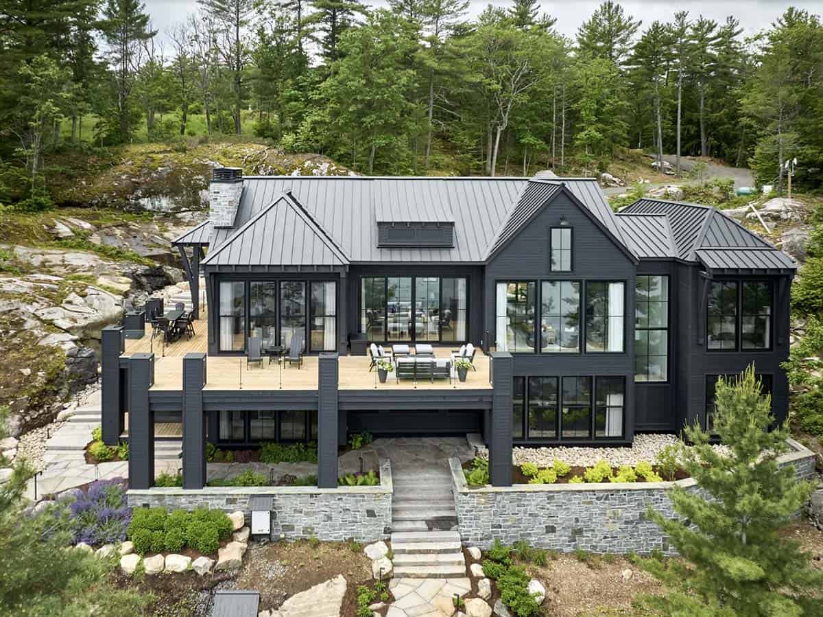 Aerial view of dark two-story cottage lake facade with large upper deck, seating areas, and stone retaining wall