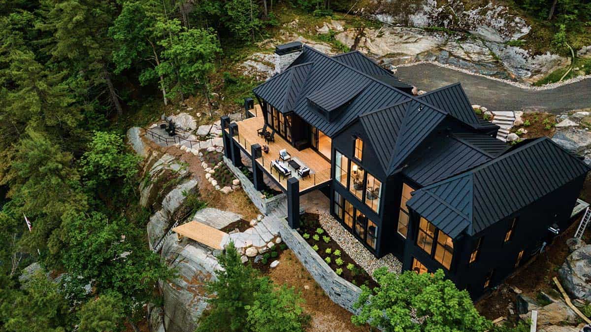 Aerial twilight view of black cottage with glowing interior lights, upper deck, and stone chimney