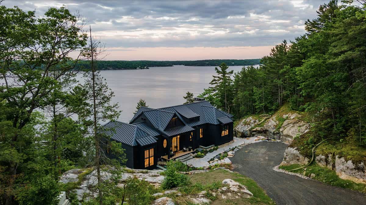 Dusk view of black cottage with warm interior glow framed by pine trees and vast lake panorama
