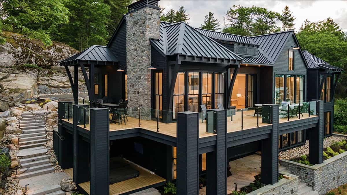 Twilight exterior of dark two-story cottage with glowing interior, stone chimney, and upper deck seating