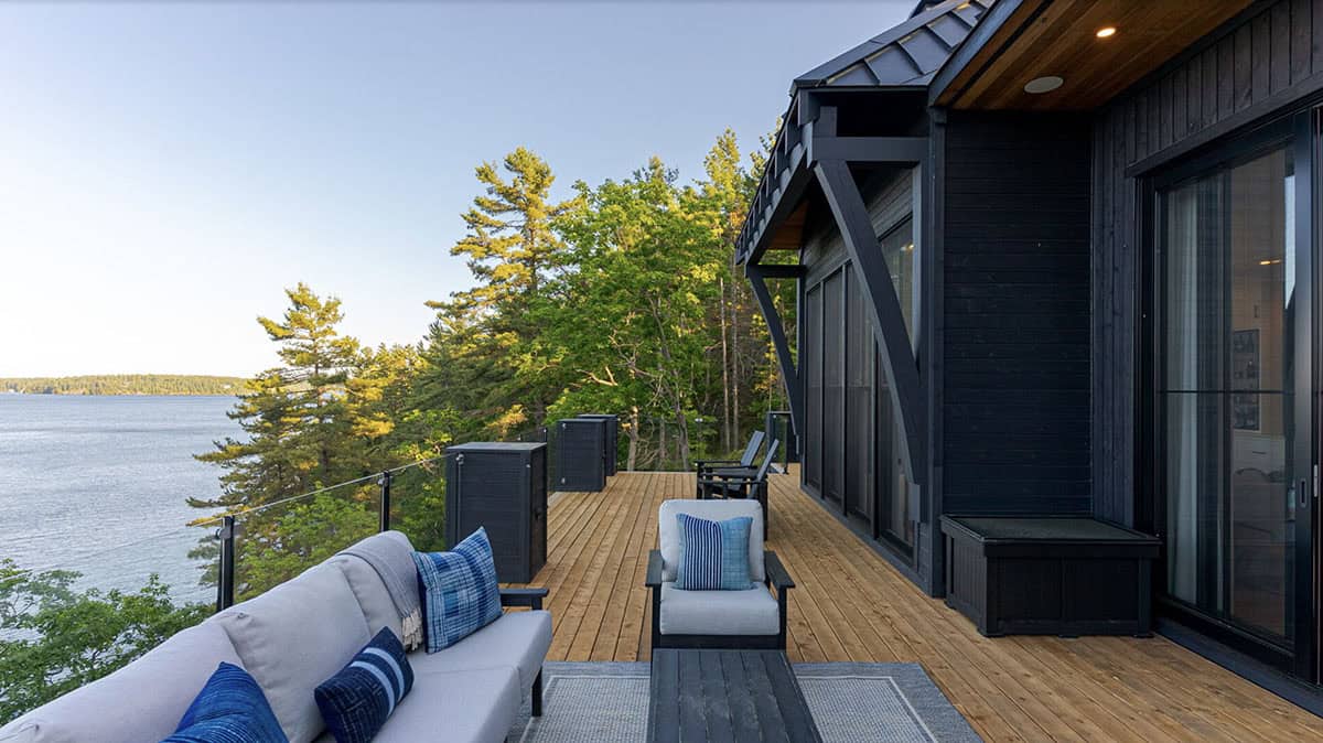 Lakeside upper deck with blue and white outdoor sofa, Muskoka chairs, and pine tree views