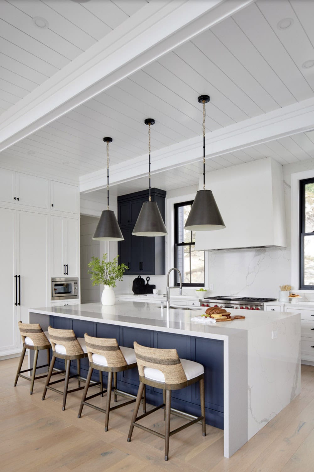 Navy blue kitchen island with woven barstools, three black cone pendants, and marble backsplash