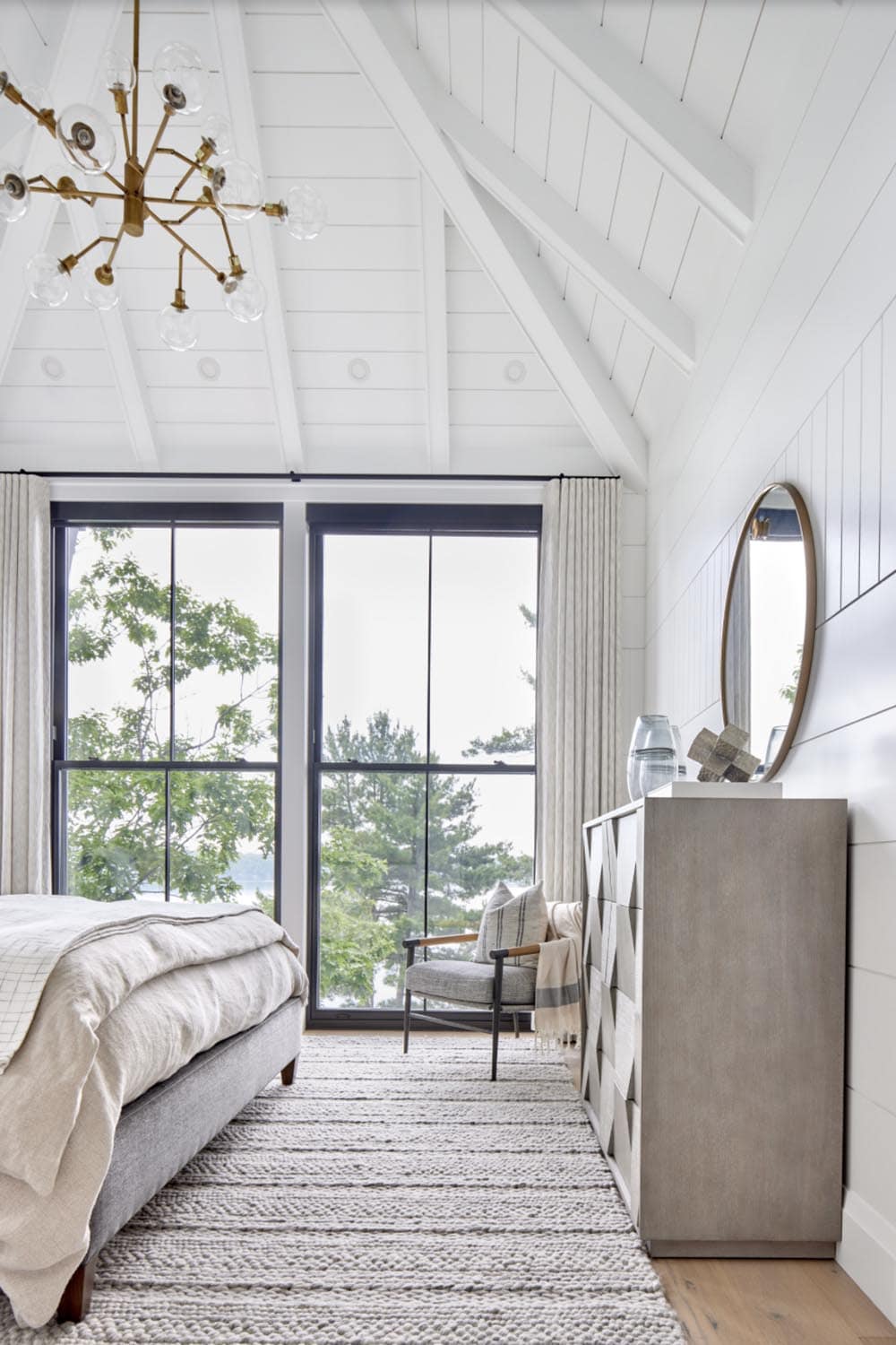 Primary bedroom corner with dresser, round brass mirror, reading chair, and black-framed lake-view windows