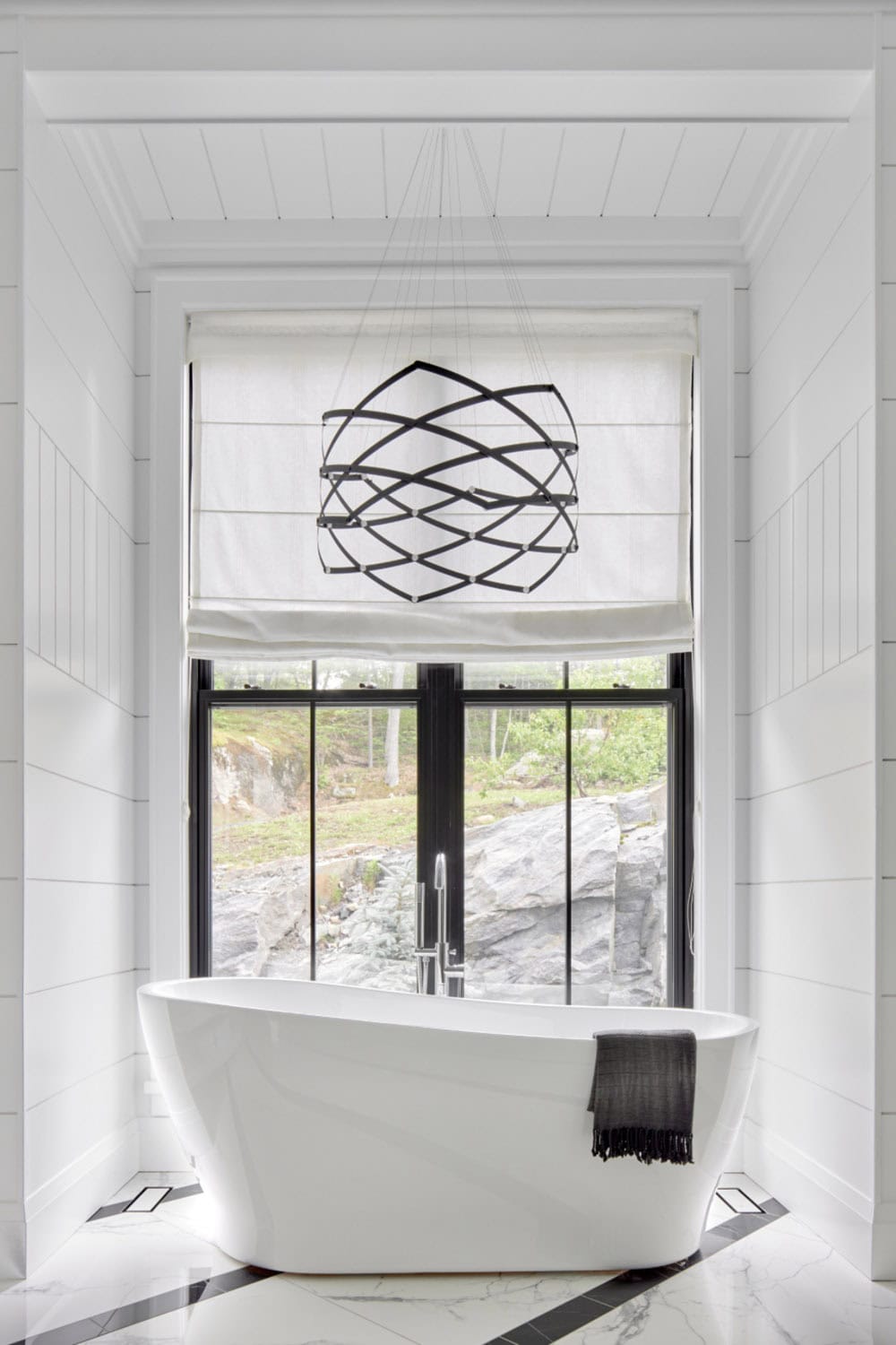 Close-up of freestanding white soaking tub centered below black geometric chandelier and large window