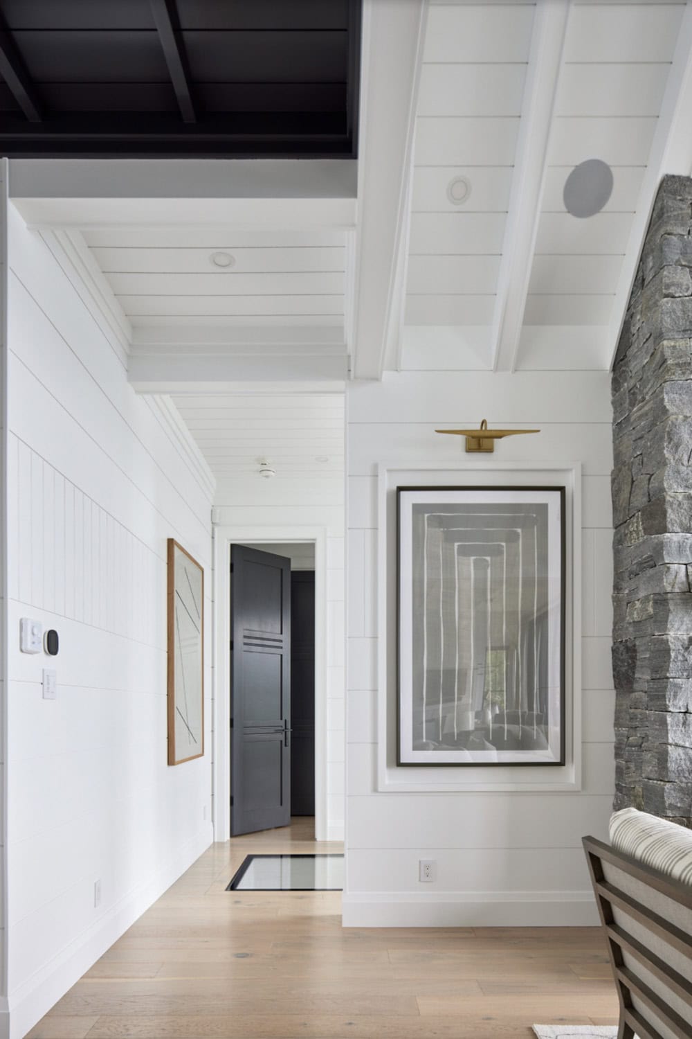Cottage hallway with white shiplap walls, abstract artwork, brass picture light, and stone fireplace edge